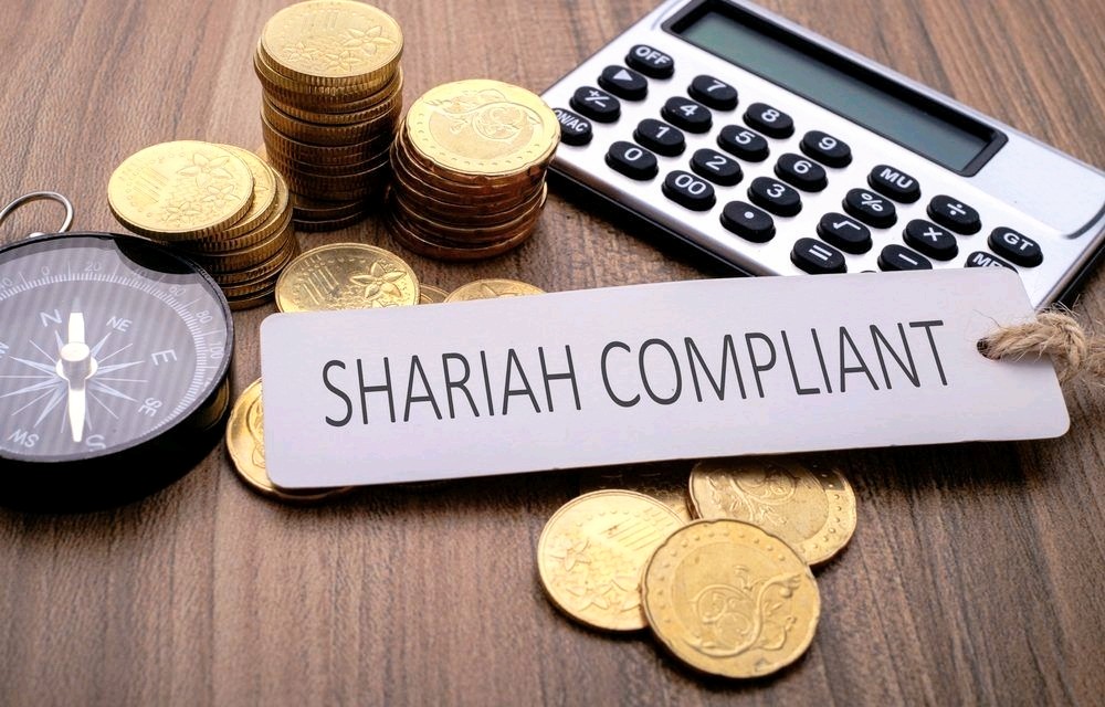 A "Shariah Compliant" sign surrounded by stacks of gold coins, a compass, and a calculator on a wooden surface.